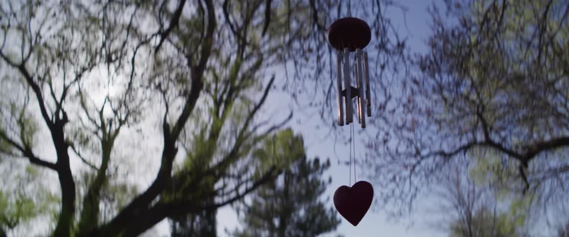 A red, heart-shaped wind chime gently sways in the breeze while hanging from a tree on a peaceful spring day