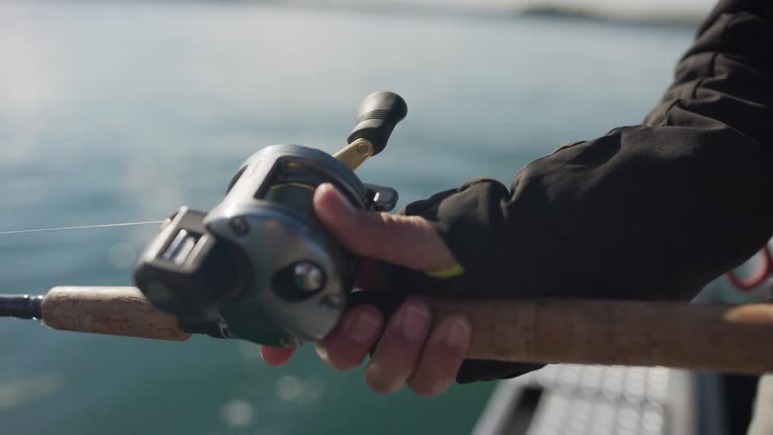 A fisherman's hands hold a baitcasting reel steady over the calm, sunlit water