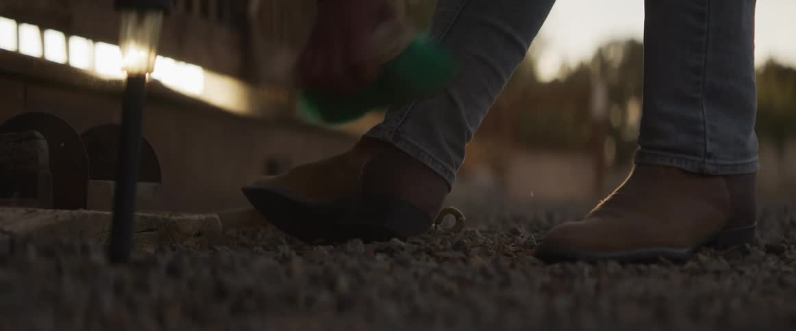A cowboy meticulously brushes the dust off his leather boots with a green brush in the warm light of sunset