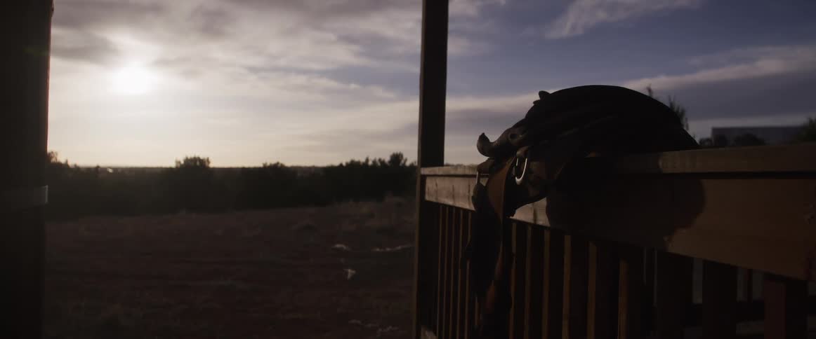 A leather horse saddle rests on a wooden porch railing at sunset, overlooking a vast and peaceful rural landscape