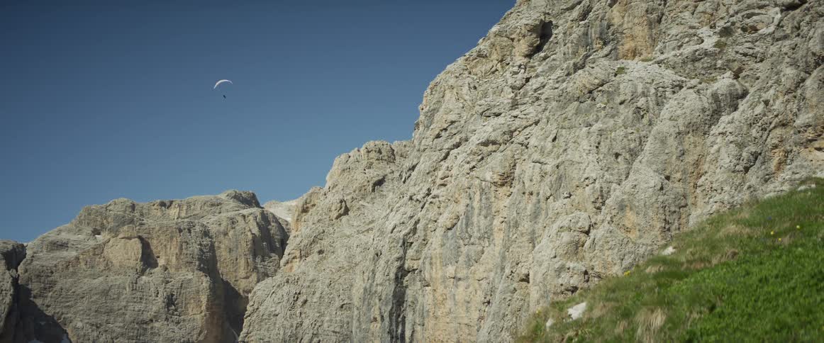 A lone paraglider navigates the air currents beside a majestic, craggy mountain range on a sunny day