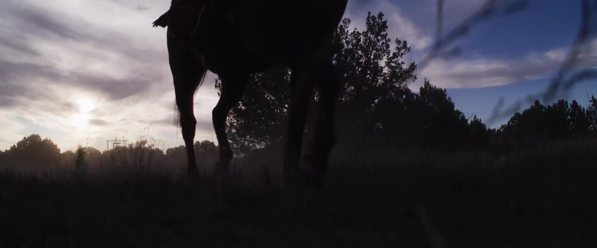 Dramatic slow-motion shot from a low angle shows two silhouetted cowboys riding horses through a dusty field at dusk