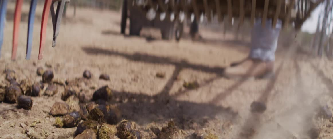 A close-up slow-motion shot shows a manure fork raking up horse droppings in a dusty paddock