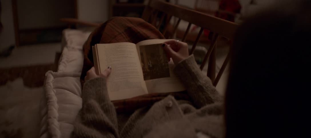 A woman cozied up on a couch reads a vintage book and finds an old photograph tucked inside before closing it