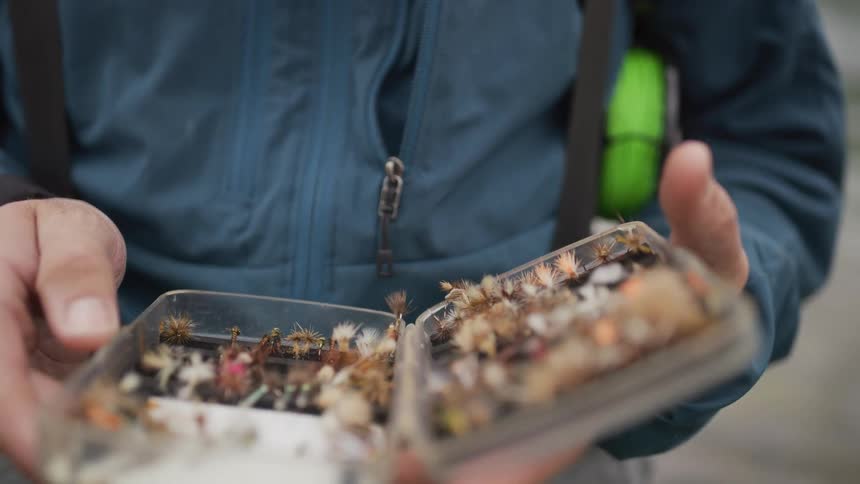 A slow-motion close-up shows a fly fisherman opening a clear tackle box to reveal a collection of colorful flies