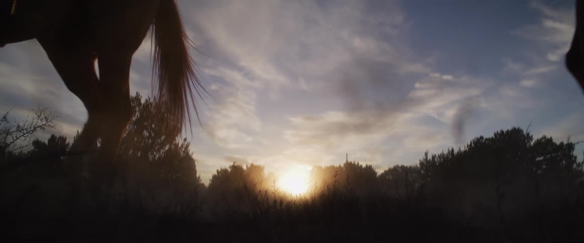A low-angle, slow-motion shot captures the silhouettes of two cowboys on horseback as they ride out of frame into the sunset