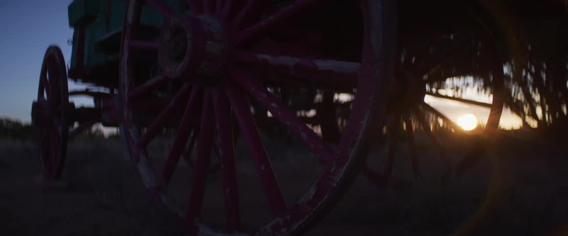 A weathered, pink wagon wheel sits still in a field as the sun sets in the background