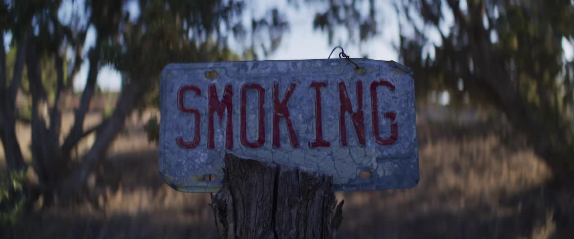 A rustic 'Smoking' sign made from an old license plate is posted on a wooden stump in a dry, rural field