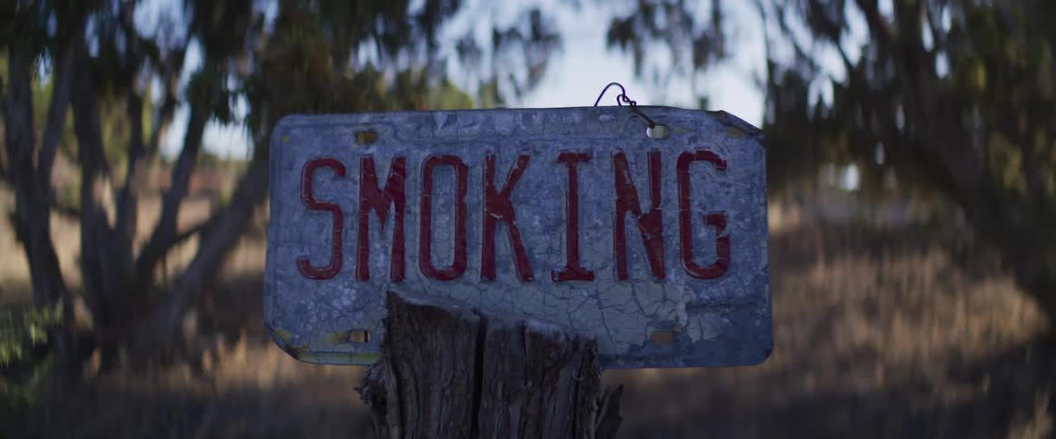 A close-up of a weathered license plate sign reading "SMOKING" hangs on a wooden post in a dry, rural landscape