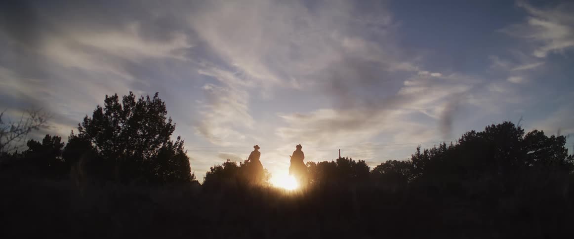 Silhouettes of two cowboys on horseback slowly ride over a hill against a stunning golden hour sunset