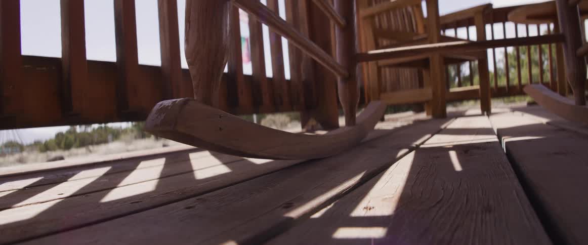 The base of a wooden rocking chair sways back and forth in slow motion on a sun-drenched wooden porch