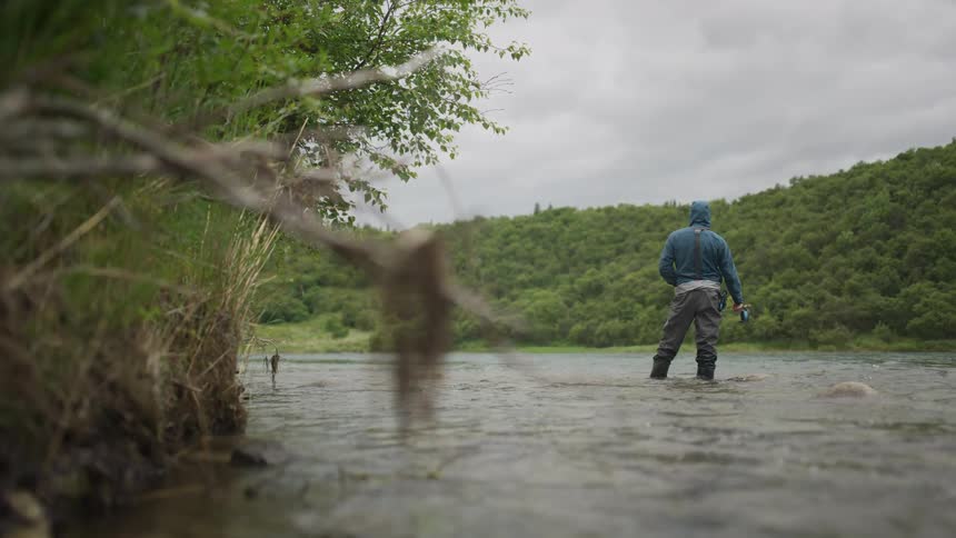 From a low water-level perspective, a lone angler in waders stands peacefully in a flowing river on a gray, overcast day