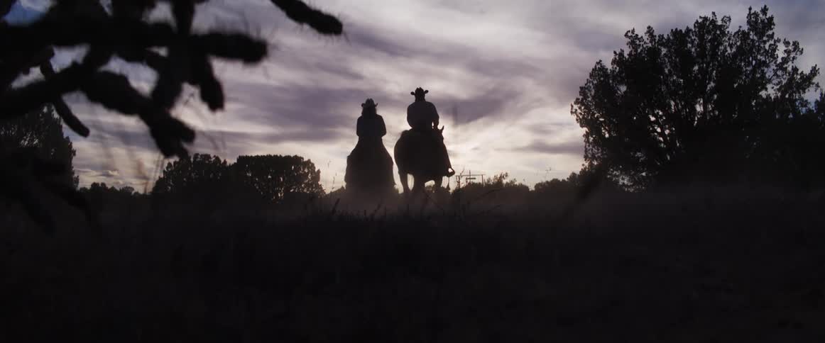 In a dramatic slow-motion shot, the silhouettes of two cowboys on horseback ride away into the dusty sunset