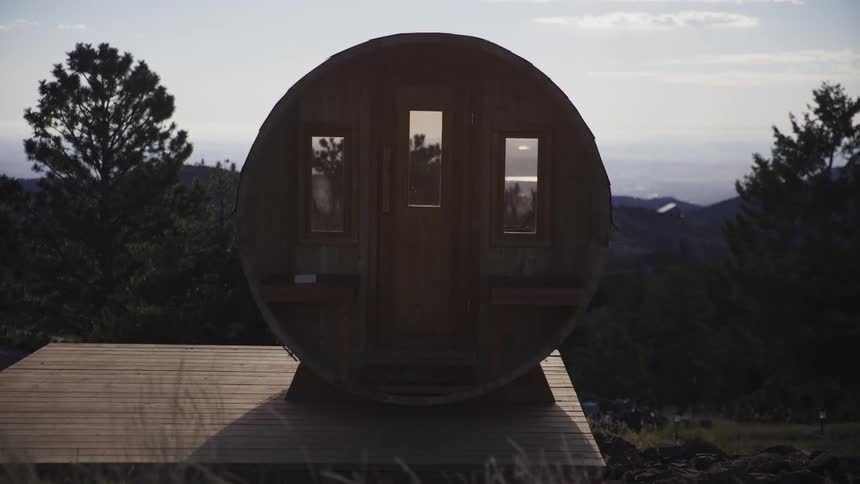 Slow dolly shot through tall grass reveals a rustic wooden barrel sauna on a mountain at dusk