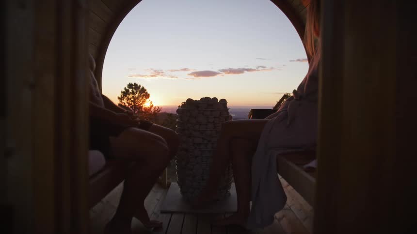 A push-in shot reveals a couple relaxing in a barrel sauna, enjoying the scenic mountain view at sunset