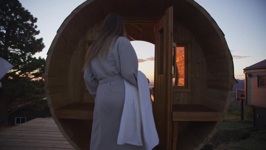 A woman in a bathrobe with a towel walks into a wooden barrel sauna on an outdoor deck during sunset in slow motion