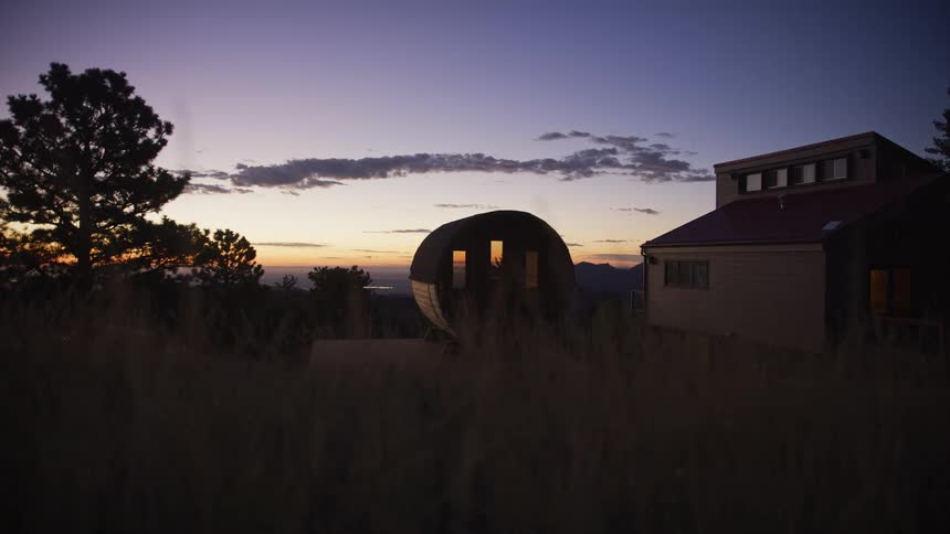 A serene twilight view of a barrel sauna steaming next to a modern house in the mountains