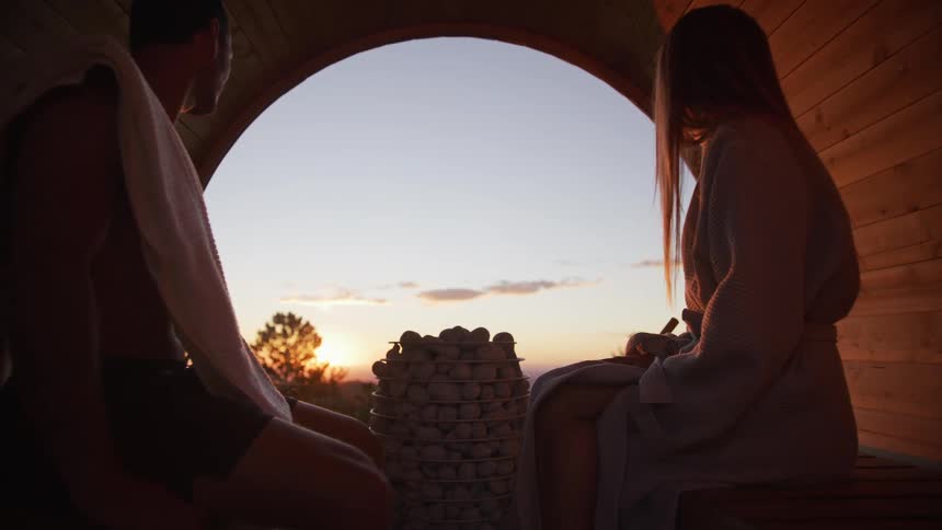 A tranquil shot reveals a couple relaxing inside a barrel sauna, enjoying the picturesque sunset view over the mountains