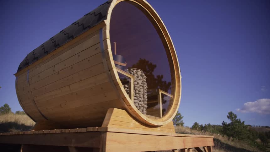 A modern wooden barrel sauna with a panoramic glass window is revealed on a sunny hillside