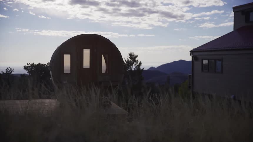A rustic barrel sauna sits in a windy, grassy meadow with scenic mountain and ocean views at golden hour