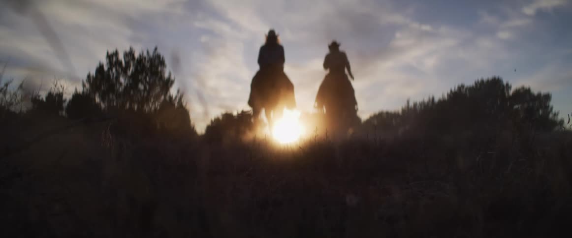 A cinematic slow-motion shot from a low angle shows two cowboys riding their horses into the golden sunset