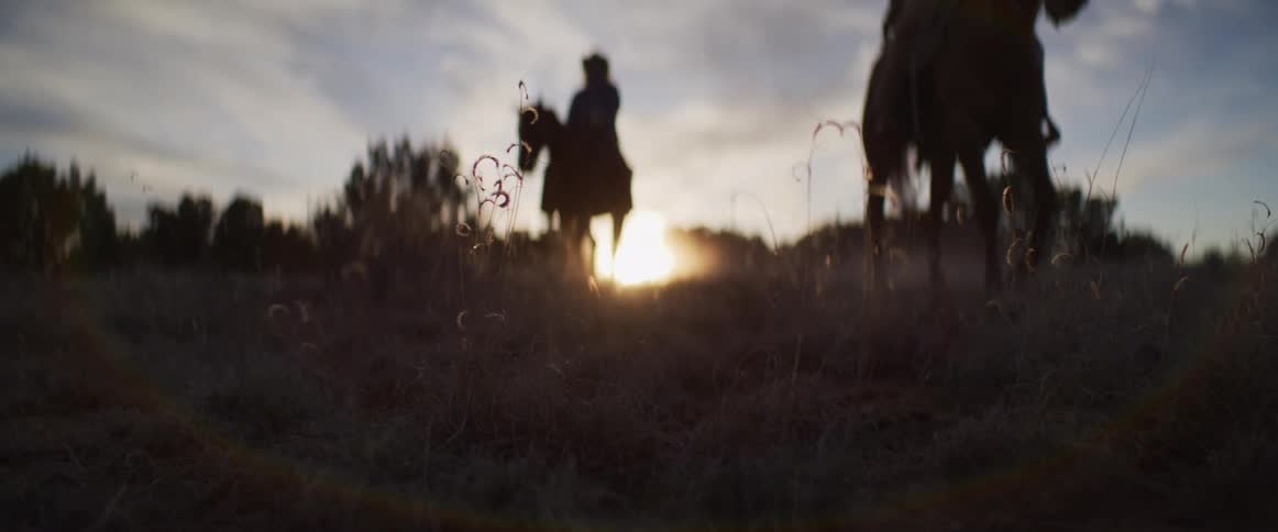 Slow-motion, low-angle shot of two silhouetted figures riding horseback through a dusty field at sunset