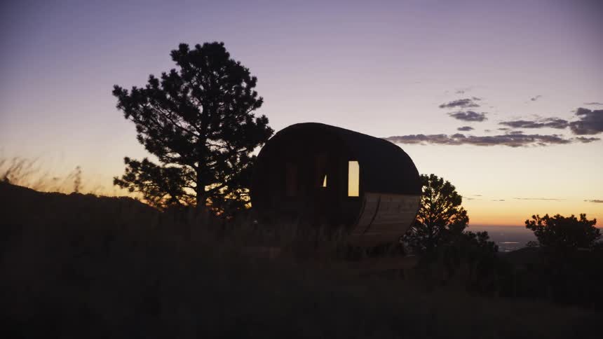A slow dolly shot reveals a cozy barrel sauna silhouetted on a hillside against a beautiful twilight sky