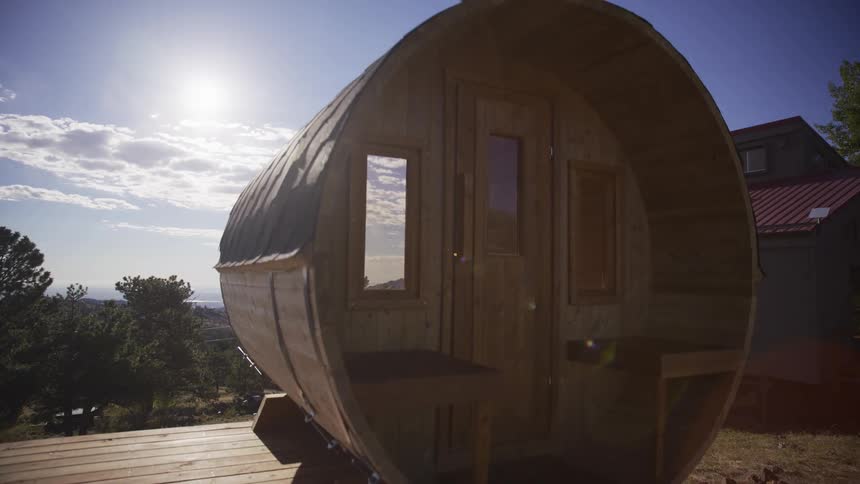 A slow and cinematic revealing dolly shot of a unique wooden barrel sauna in a beautiful sunlit outdoor setting