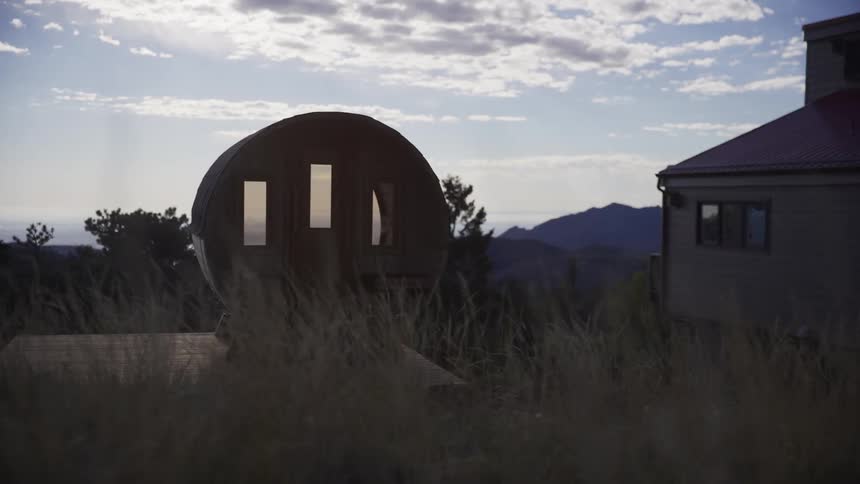 A slow pull-back shot reveals a rustic barrel sauna nestled in a grassy field next to a house at dusk