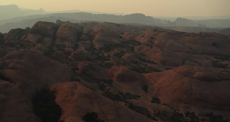 A static wide shot captures the vast, hazy red rock landscape of the desert during a golden hour sunset