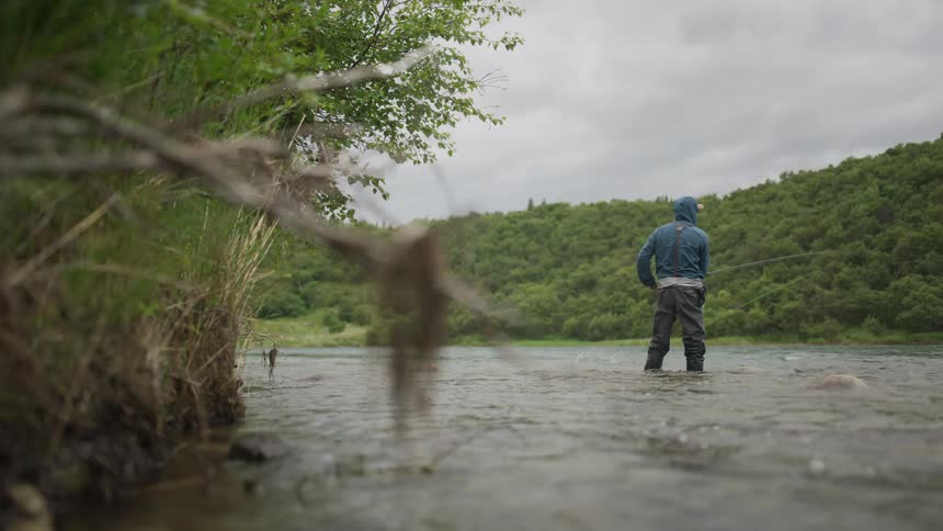 A lone fly fisherman wearing waders and a hood stands in a flowing river casting his line on an overcast day in the wilderness