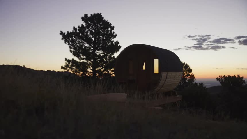 A beautiful wooden barrel sauna is silhouetted against a colorful sunset sky on a remote mountainside