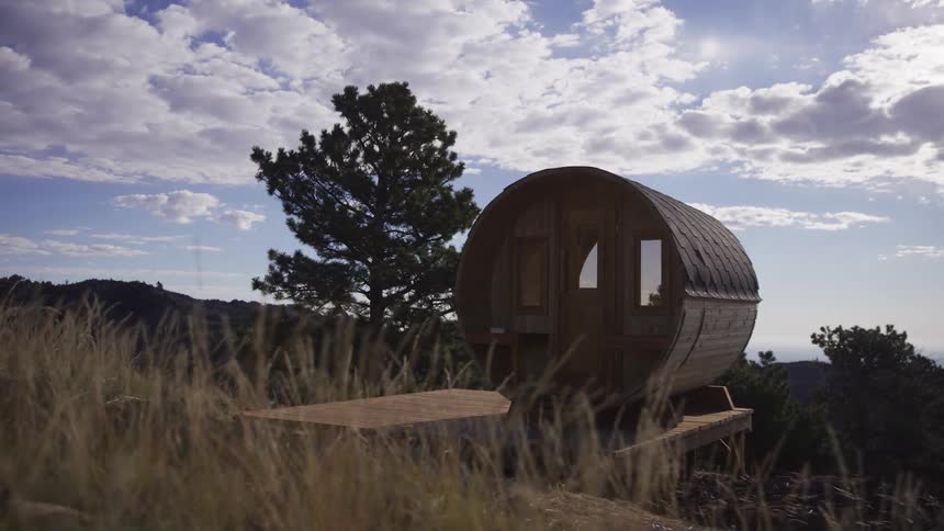 A rustic wooden barrel sauna sits on a peaceful hillside with tall grass swaying in the breeze