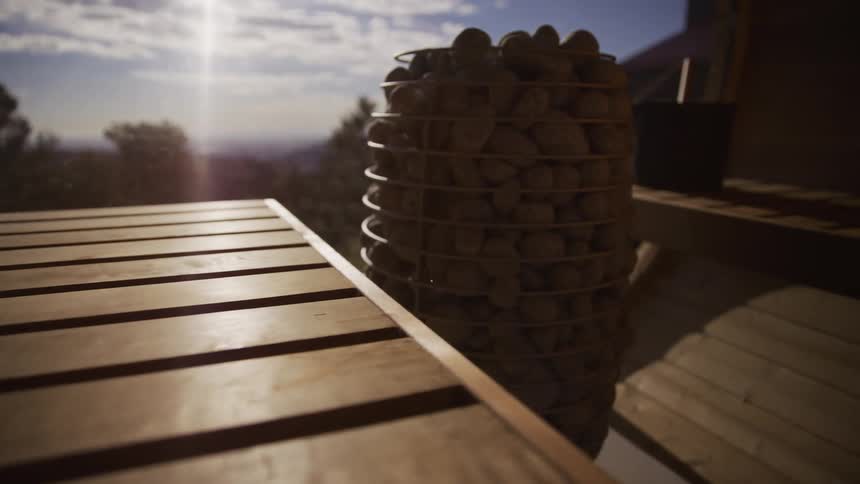 A slow pan reveals the warm interior of a wooden sauna with a stone heater as sunlight streams through a window overlooking a scenic landscape