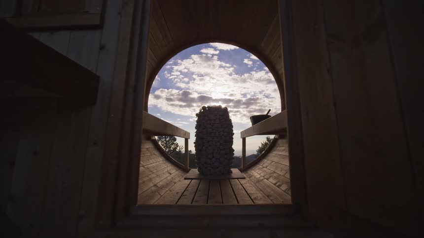 Slow push-in shot revealing the beautiful interior of a modern wooden barrel sauna with a large circular window framing a cloudy sky