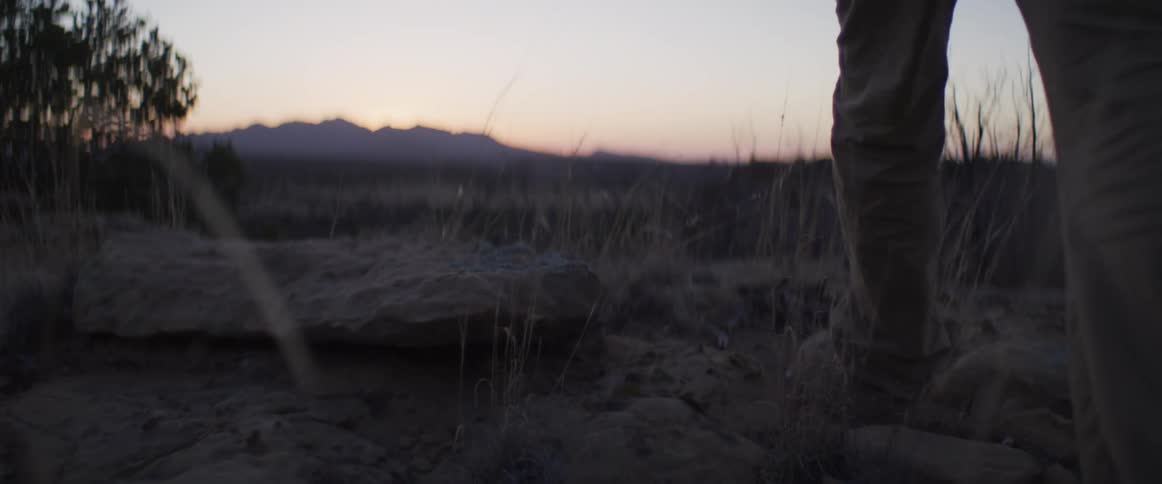 A slow-motion, low-angle shot of a person's legs and boots hiking through a desert landscape towards distant mountains at sunset