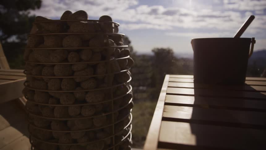 Slow-motion push-in shot of a hot sauna heater and wooden bucket in a barrel sauna with a sunny nature view