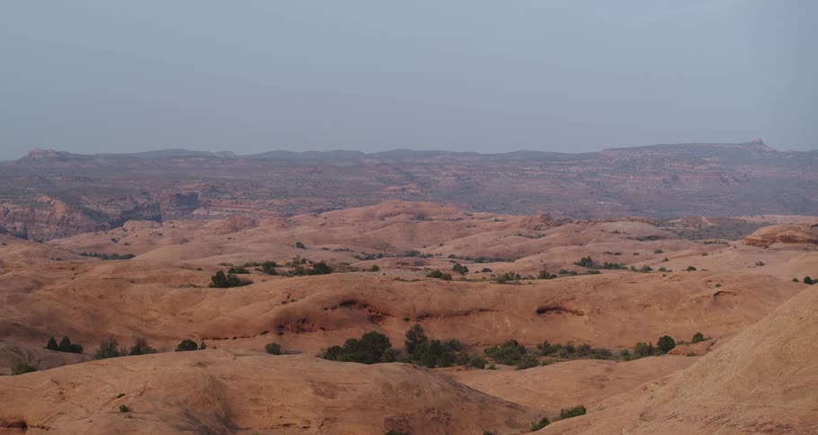 A slow pan reveals the expansive, undulating red rock formations of a desert landscape under a hazy sky