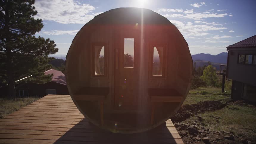 A barrel sauna sits on a wooden deck in the mountains as the camera pulls back and the sun flares