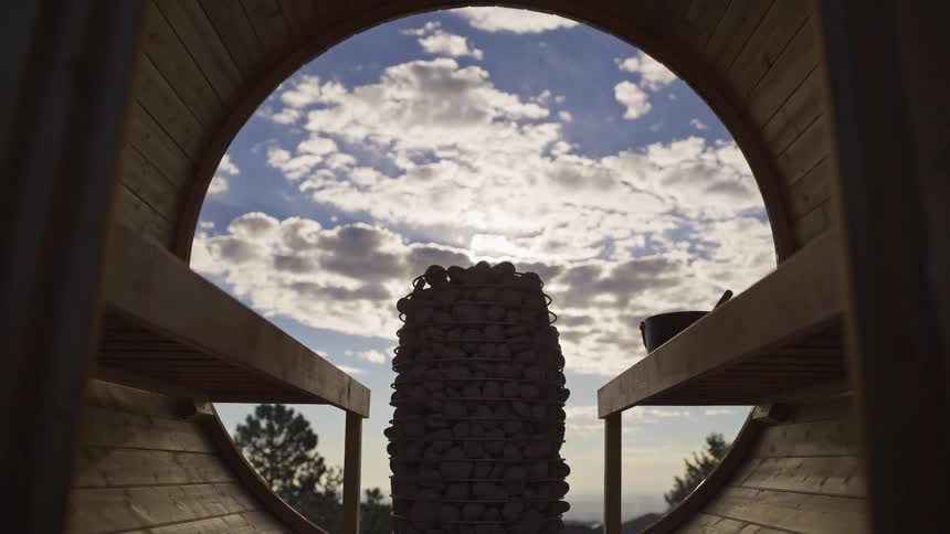 A serene pull-out shot from inside a wooden barrel sauna reveals a stone heater and a beautiful view of the sky through a large circular window