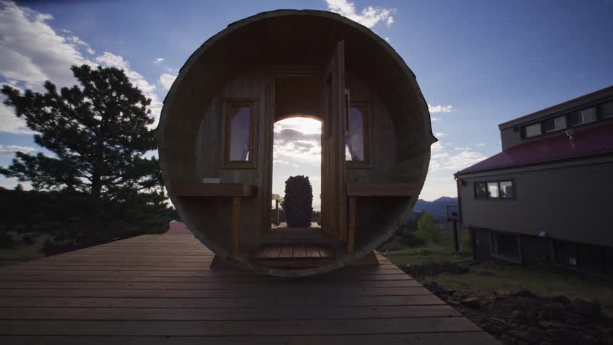 A unique wooden barrel sauna sits on an outdoor deck as the sun glows through its open doors