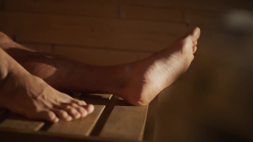 Close-up slow-motion shot of a man's feet relaxing on a wooden bench inside a sauna