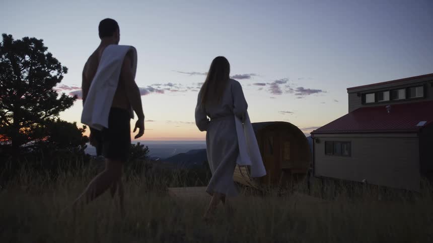 A couple walks toward a scenic mountainside barrel sauna for a relaxing evening at sunset