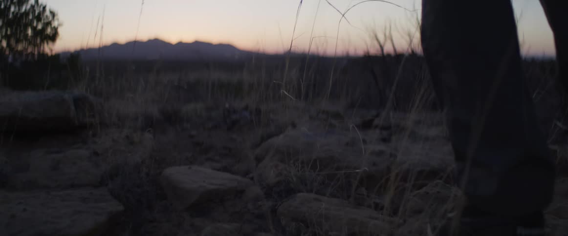 A low-angle slow-motion shot of a person's feet hiking through a rocky, grassy desert landscape at sunset