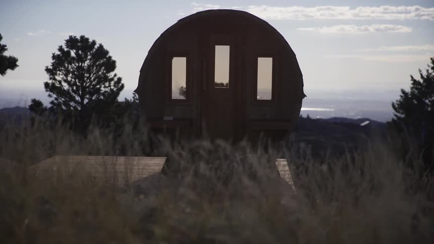 Slow dolly shot through tall grass reveals a rustic wooden barrel sauna on a hillside overlooking a valley at sunrise