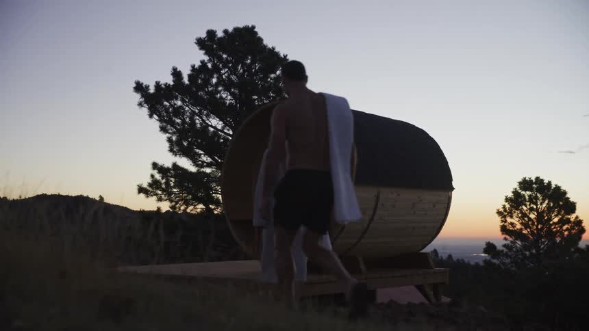 A silhouetted couple walks toward a barrel sauna on a tranquil mountainside during a beautiful sunset