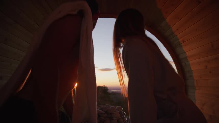 A couple relaxes together in a cozy barrel sauna, watching a beautiful sunset over the distant hills