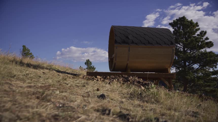 A low-angle slider shot moves toward a wooden barrel sauna perched on a grassy hillside under a blue sky