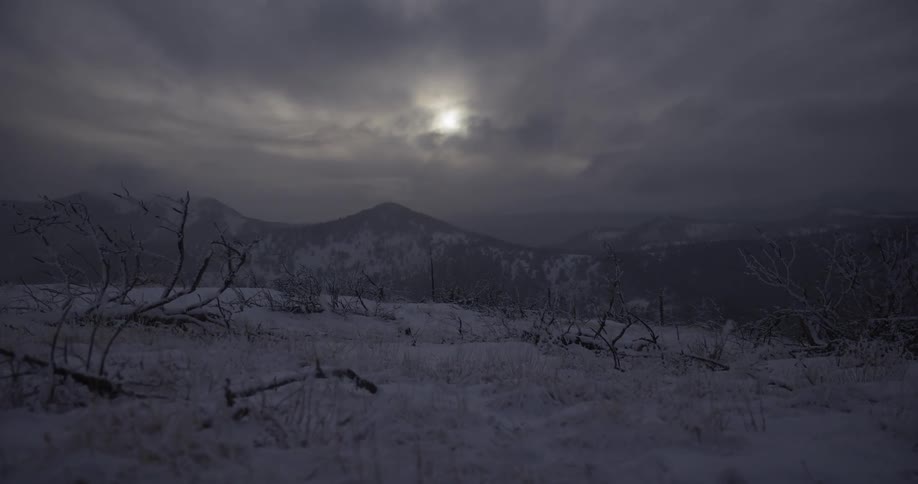 A stark, snow-covered mountain landscape rests under a dark, moody sky with a faint sun breaking through the clouds
