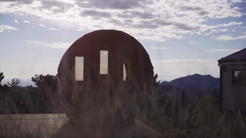 A slow pull-out shot reveals a unique wooden barrel sauna overlooking a majestic mountain range and distant city at golden hour